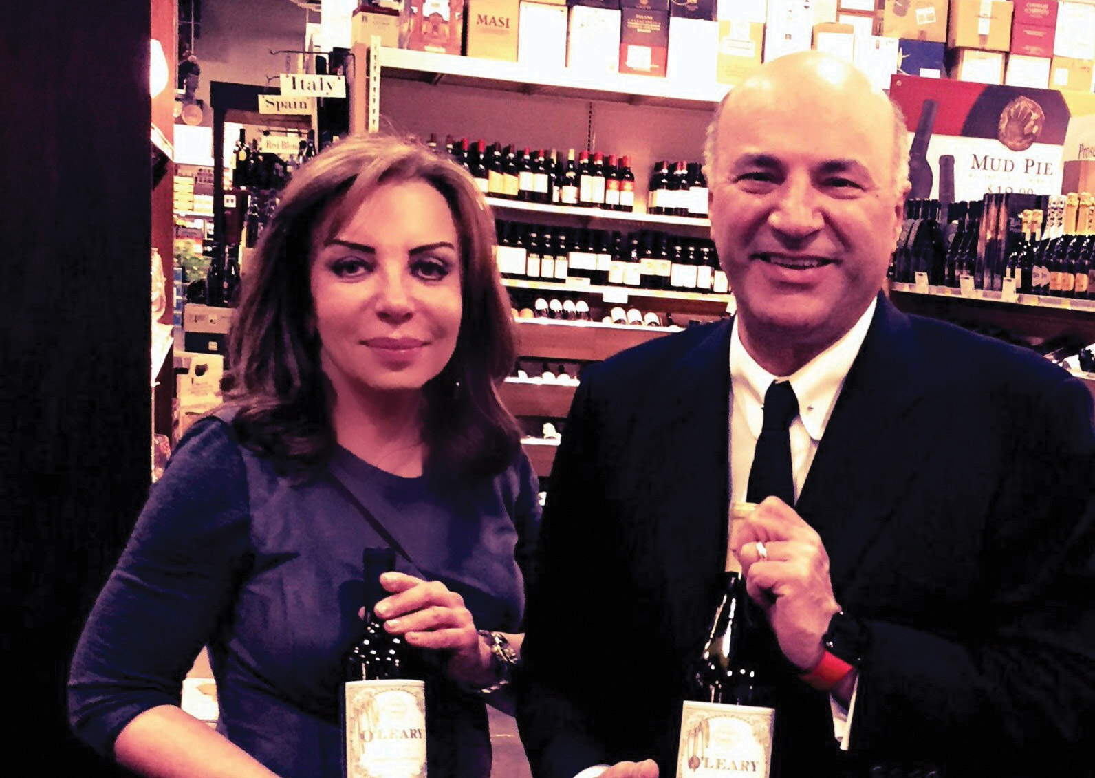 Anne Marie Harrison with Kevin O'Leary standing in her wine store each holding a bottle of wine.