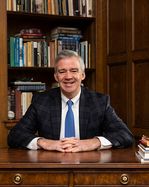 President Carey sits at his desk with a shelf full of books behind him